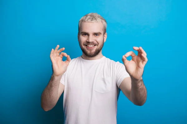Homem com barba e cabelo loiro está posando em uma parede de estúdio azul gesticulando o sinal ok — Fotografia de Stock