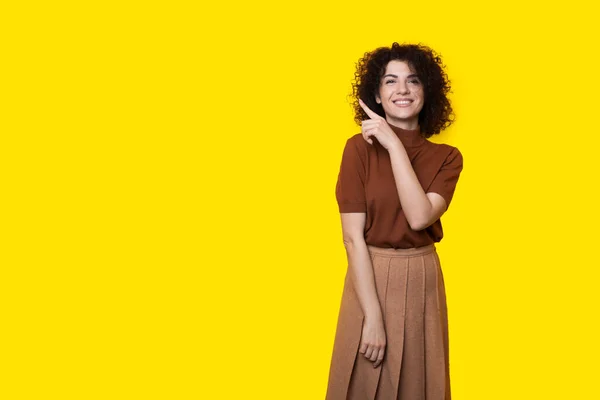 La signora dai capelli ricci con un bel sorriso sta pubblicizzando qualcosa su un muro giallo dello studio indicandolo. — Foto Stock