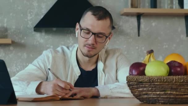 Caucásico hombre concentrado está escribiendo algo en un copybook mientras trabaja en la tableta desde casa — Vídeo de stock