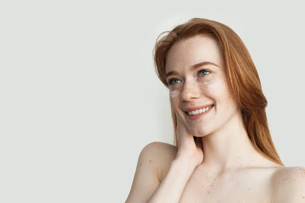 Vrouw met sproeten en rood haar poseert op een witte studio muur van toepassing op een haar gezicht anti aging crème — Stockfoto