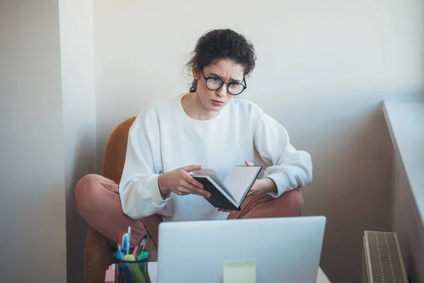 Koncentrerad kvinna tar online lektioner på laptop med hjälp av en bok och bär glasögon — Stockfoto