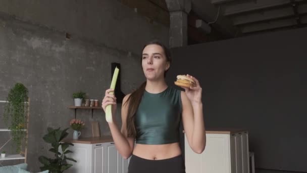Brunett kvinna äter en hamburgare medan du håller en naturlig purjolök i handen hemma — Stockvideo