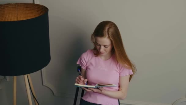 Caucasian ginger lady is smiling while doing homework and writing in a copybook — Vídeo de stock