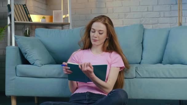 A vörös nő könyvet olvas a padlón, miközben tanul valamit a padlón ülve. — Stock videók