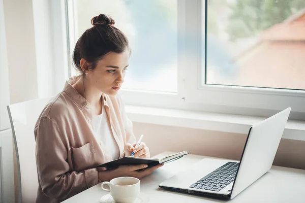 Brunette woman is writing in a copybook something while looking at the laptop screen during workday at the office — Stock Photo, Image