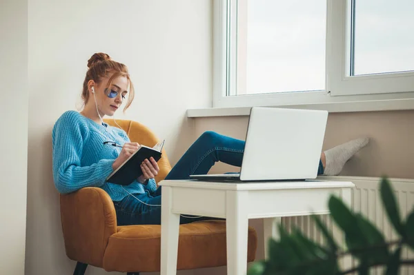 Mujer jengibre con pecas está usando parches para los ojos de hidrogel mientras está acostado en el sillón y haciendo la tarea en el ordenador portátil — Foto de Stock