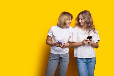 Çekici sarışın kardeşler birbirlerine gülümsüyor sarı bir duvarda poz veriyorlar ve cep telefonlarıyla sohbet ediyorlar.