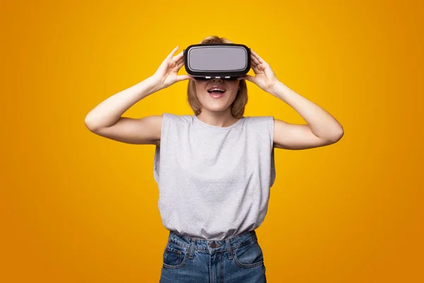 Mulher adorável está sorrindo ao usar um fone de ouvido realidade virtual ficando surpreso em uma parede de estúdio amarelo — Fotografia de Stock