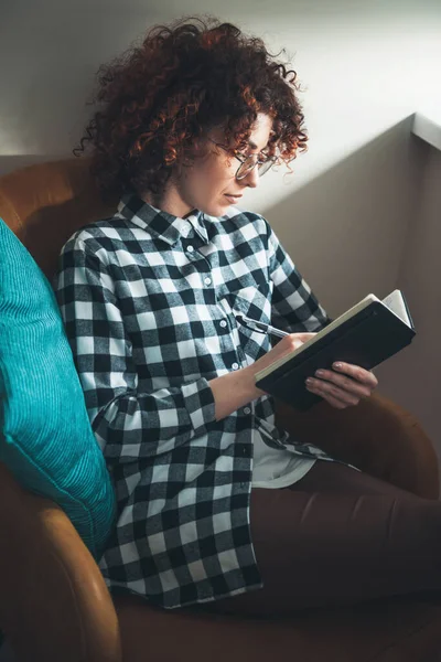 En vacker kvinna med lockigt hår skriver i en bok när hon ligger i en fåtölj hemma i glasögon. — Stockfoto