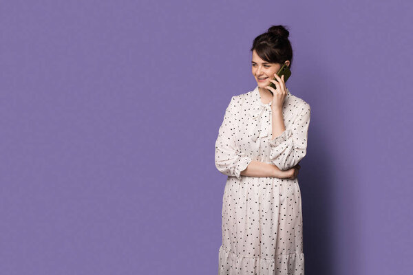 Brunette woman is smiling while posing in a dress on a blue wall with free space while having a phone conversation