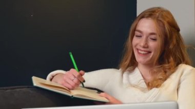 Kızıl kadın evde bir kanepede çalışıyor bir şeyler yazıyor ve gülümsüyor.