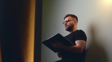 Sakallı beyaz bir öğretmen elinde bir kitapla evde ders veriyor ve kameraya konuşuyor.