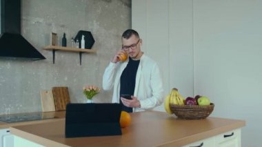 Kafkasyalı bir adam iş görüşmesi yaparken mutfakta bir tablet kullanıyor ve portakal yiyor.