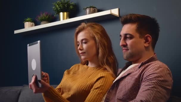 Mujer adorable con el pelo de jengibre está sentado cerca de su amante en un sofá usando una tableta — Vídeo de stock