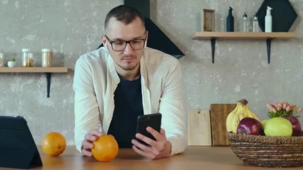 Καυκάσιος άντρας χρησιμοποιεί ένα τηλέφωνο ενώ κρατάει ένα πορτοκάλι στην κουζίνα και χρησιμοποιεί μια ταμπλέτα για επαγγελματικές συμφωνίες. — Αρχείο Βίντεο