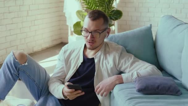 Jovem homem de negócios está usando um telefone enquanto pensa e sentado no chão perto do sofá — Vídeo de Stock
