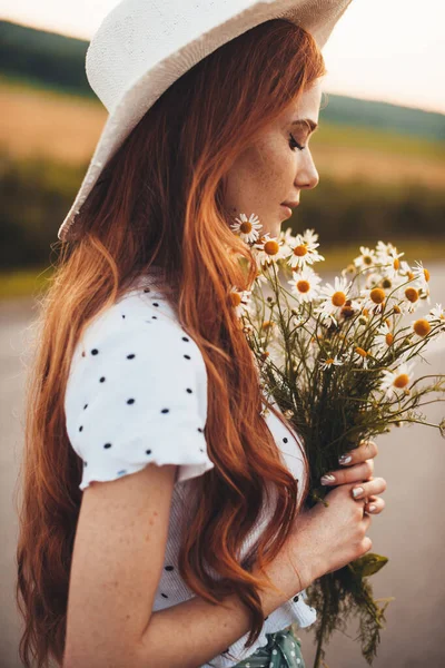 Close up photo of a ginger woman with freckles holding a bunch of flowers wearing a hat in the field