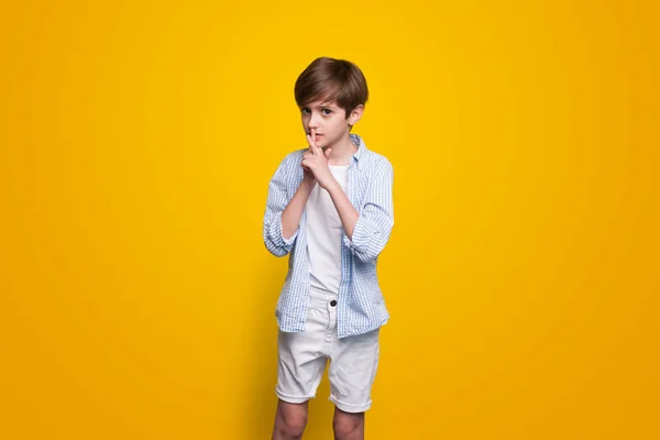 Menino adorável está gesticulando o sinal de silêncio posando em roupas brancas em uma parede de estúdio amarelo — Fotografia de Stock