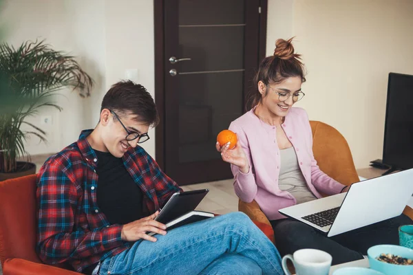 Genç iş çifti evde tablet ve dizüstü bilgisayarla çalışıyor gülümsüyor ve ellerinde meyve tutuyorlar. — Stok fotoğraf