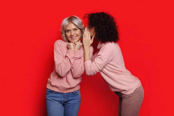 Roter Hintergrund. Schwestern tratschen. Flüstern am Ohr ein Geheimnis. — Stockfoto