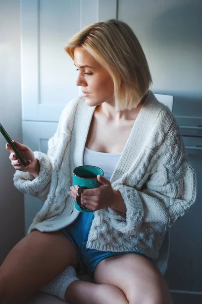 Hermosa mujer rubia con teléfono en la cocina. Persona que bebe café. Comunicación móvil. Hogar inteligente. — Foto de Stock