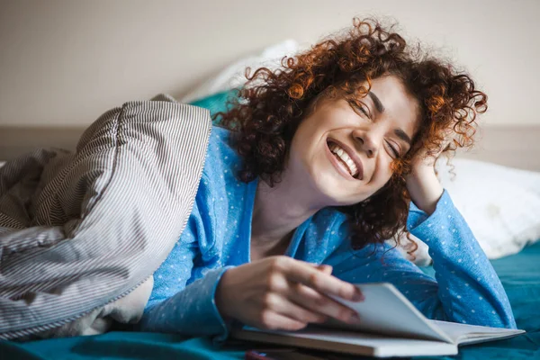 Wanita keriting mengenakan piyama di tempat tidur. Foto pendidikan dari buku bacaan seseorang. Konsep homeschooling. — Stok Foto