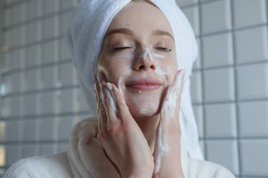 Kadın yüzünü nazikçe yıkıyor, rahat bir kadın sabah tenini köpükle ve yumuşak bir gülümsemeyle yıkıyor.