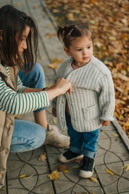 Yürek ısıtan bir sahne, gülen çocuğunu canlı düşen yaprakların arasında dikkatlice giydiren sevgi dolu bir anneyi tasvir eder.