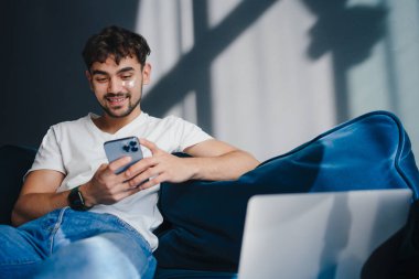 Genç bir adam, şık, modern bir iç mekanda akıllı telefonunu yanında bir dizüstü bilgisayarla kanepeye uzanır.