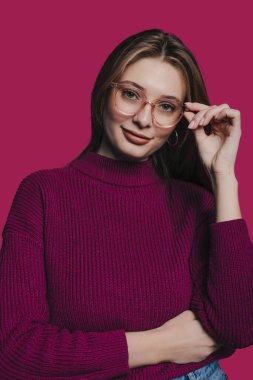 Şık genç bir kadın kendine güvenen pembe bir süveter ve şık gözlüklerle poz verir.