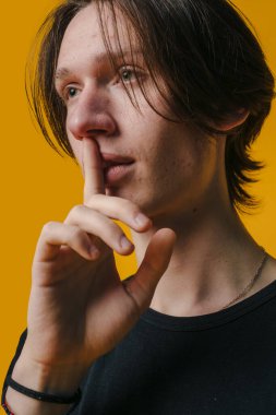 Genç bir adam, saçları uzun ve dalgalı, canlı ve parlak sarı bir zemin karşısında sessizlik için jest yapıyor.