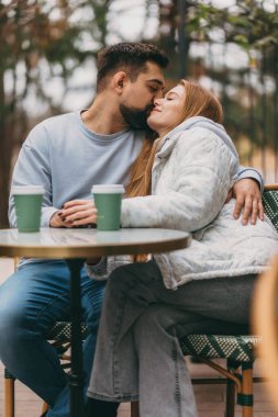Kahve fincanlarının üzerinde öpüşen çift, rahat teras sahnesi, sıcak sohbet, masadaki eller, gündelik sonbahar katmanları.