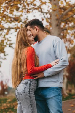 Romantik park resminde poz veren çift, kadın gülümserken erkeğin alnını öpen adam, şık katmanlar, arka planda düşen yapraklar.