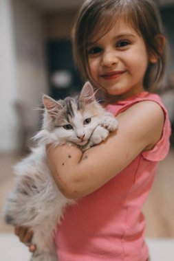 Genç bir kız kucağında kedi yavrusuyla gülümsüyor. Kutlama sahnesi. Parlak bir ifade, rahat bir oturma odası.,