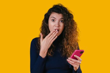 Kafkasyalı kadın telefona şaşırmış, ağzı kapalı pembe akıllı telefonu koyu sarı renkte okurken.