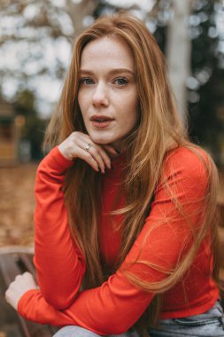 Kırmızı kazaklı, düşünceli, beyaz bir kadın. Elinde çene, sonbahar yapraklı açık hava portresi.
