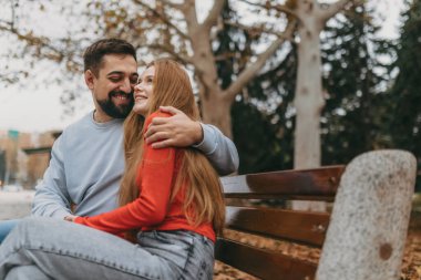 Kafkasyalı çift bankta gülerek neşeli bir sonbahar gezintisinin tadını çıkarıyor, kızıl saçlı kadın sakallı kadına yaslanıyor.