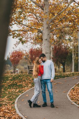 Kafkasyalı çift, sonbahar ağaçlarının altında kıvrımlı kaldırım yolunda yürüyor, nazikçe sarılıyor ve gevşek sohbetler ediyor.