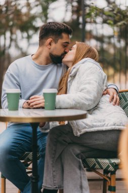 Kafeteryada öpüşen beyaz bir çift, yemek fincanları masada, hafif kahkahalar, sıradan ceketler, samimi anlar.