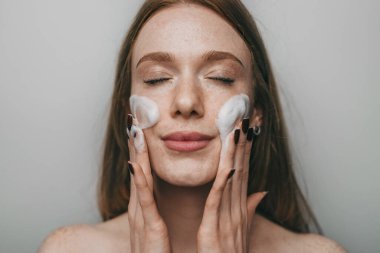 Yüzü köpükle temizleyen beyaz kadın, gözleri kapalı, yanakları okşayan, yumuşak doğal ışık, samimi.