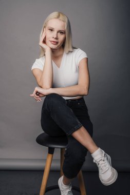 Beyaz kadın tabureye oturmuş, düşünceli bir ifade takınıyor ve tarafsız stüdyoda rahat bir poz veriyor, sarı saçlı, beyaz tenli.