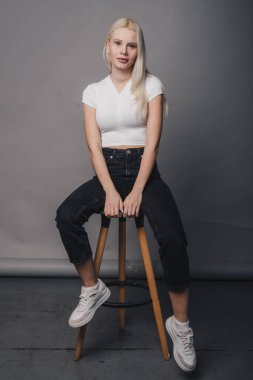 Tarafsız kadın, tahta tabureye oturmuş tişört ve siyah pantolon giyerek gri zemine yaslanmış.