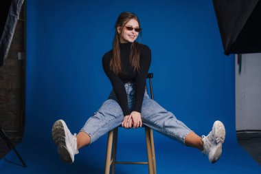 Güneş gözlüğü ve balıkçı yaka giyen, ayakları açık, rahat poz veren stüdyo taburesinde kendine güvenen bir kadın.