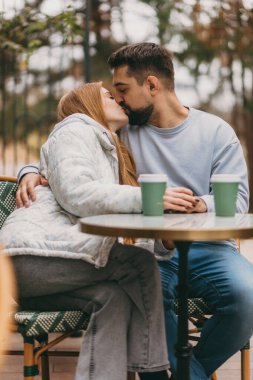 Sonbaharda açık kafede öpüşen çift, masada iki kahve fincanı, sıcak kazaklar, yakın sarılma ve yumuşak