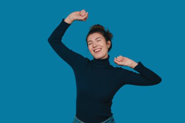 Gülümseyen kadın gözleri kapalı mavi arka planda kollarını açmış, balıkçı yaka ve kot pantolon giyiyor, rahatlamış.