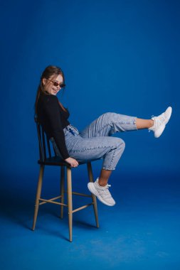 Taburede poz veren genç bir kadın, güneş gözlüğü ve kareli pantolon, siyah ceket ve beyaz spor ayakkabı giyiyor. mavi