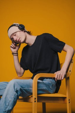 Beyaz adam, kulaklıkları ahşap koltuğa oturmuş siyah tişörtlü açık renk kot pantolonlara takmış, rahatlamış bir şekilde dinliyor.