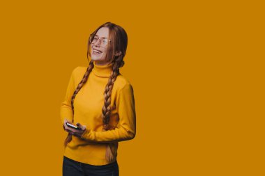 Sarı kazaklı kadın telefona bakarken gülüyordu, örgülü saçlar çerçeveli gülen yüz ve sıcak hardal arka planı.