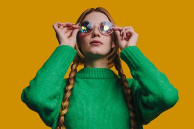 Yeşil süveter giyen genç bir kadın yansıtıcı güneş gözlüğü takıyor, örgülü örgü örüyor, canlı hardala karşı kendine güvenen bakışlar atıyor.