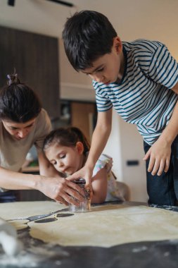 Kardeş takımı un tezgahtaki kurabiyeleri süslüyor, kız ve oğlan şekillere odaklanıyor, ebeveyn asistanı...